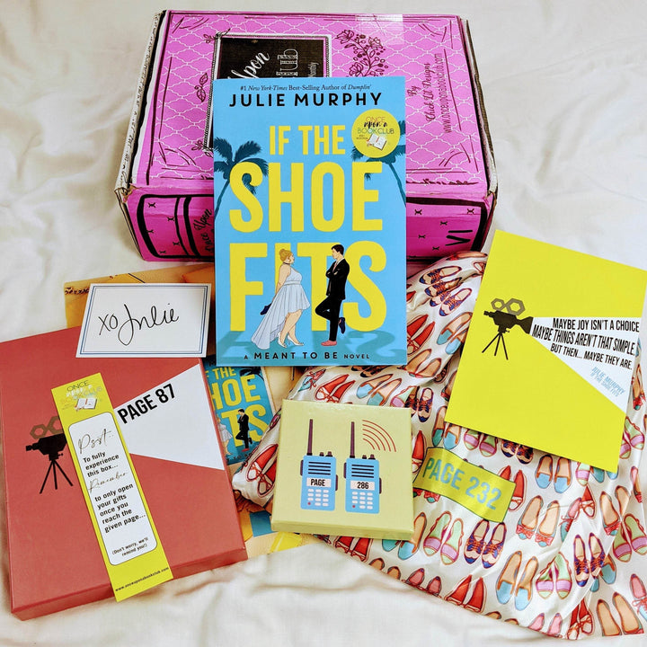 A paperback edition of If the Shoe Fits leans against a pink Once Upon a Book Club box. In front of the book are a signature card, bookmark, orange box, yellow square box, quote card, and white drawstring bag. The boxes and bags all have page numbers.