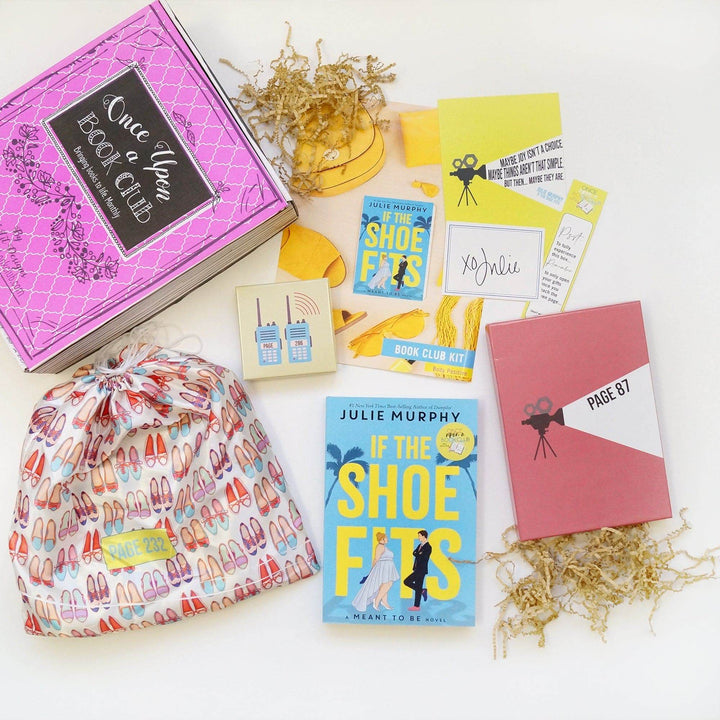 A pink Once Upon a Book Club box is at the top left of the image. In front of the box are a white drawstring bag with a pattern of shoes, yellow square box, bookclub kit, quote card, signature card, bookmark, paperback edition of If the Shoe Fits, and pink box. The boxes and bags all have page numbers.