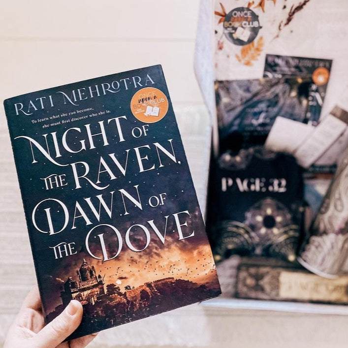 A white hand holds up a hardcover edition of Night of the Raven, Dawn of the Dove in front of a blurred background of an open box with wrapped gifts inside