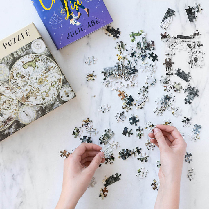 a pair of white hands hold puzzle pieces. There are puzzle pieces scattered across a marble background next to a box labeled puzzle and a hardcover edition of The Charmed List