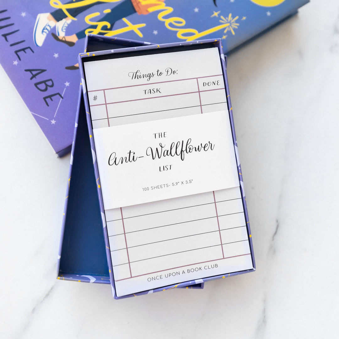 a white notepad labeled Things to Do with a white label across the center that says The Anti-Wallflower List in a box leaning against a hardcover edition of The Charmed List