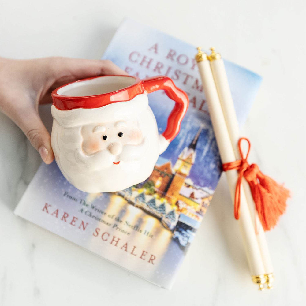 a white hand holds a mug shaped like Santa in front of a hardcover edition of A Royal Christmas Fairy Tale and a scroll tied together with an orange tassel
