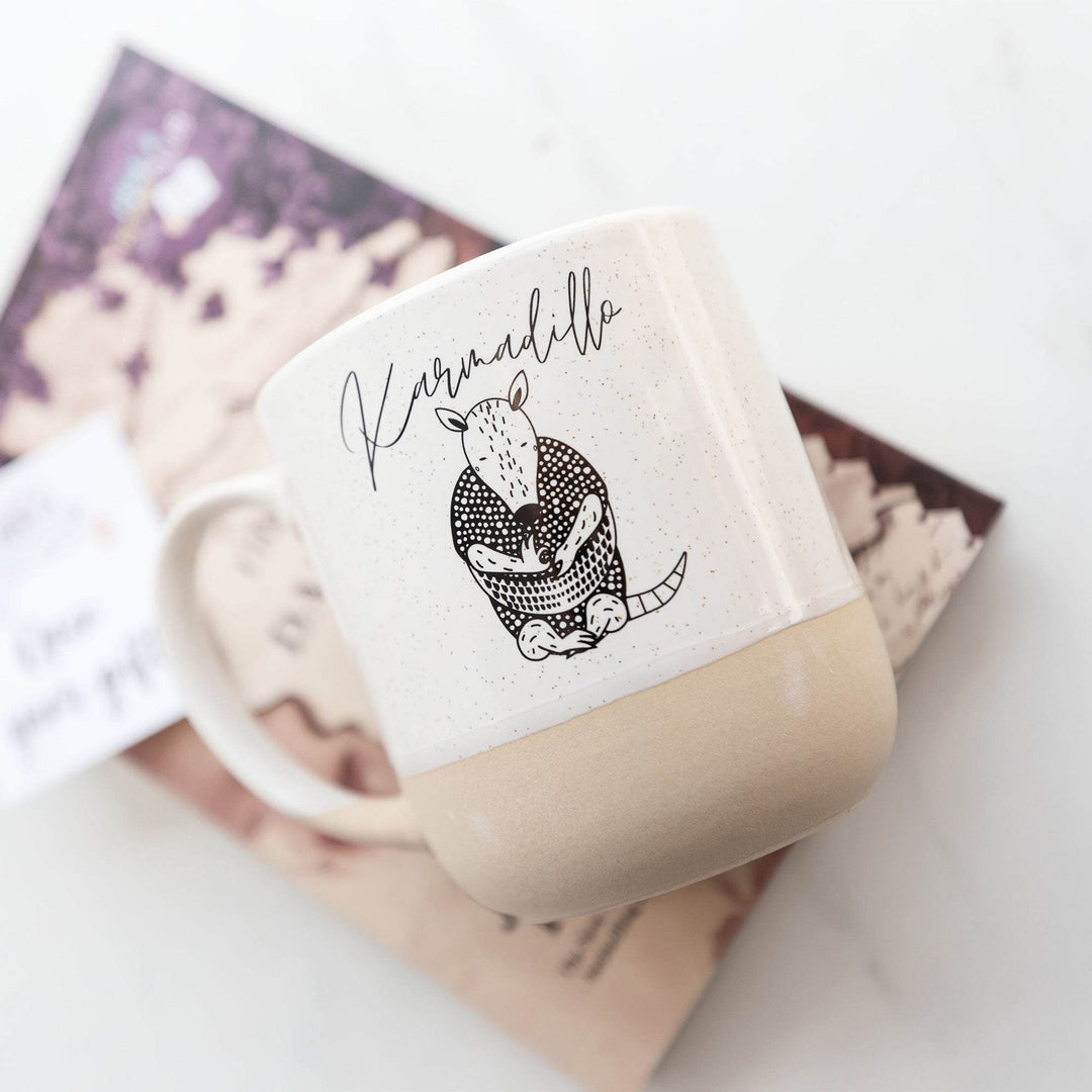 a white mug labeled "Karmadillo" with an image of an armadillo lays on a hardcover special edition of The Many Daughters of Afong Moy 