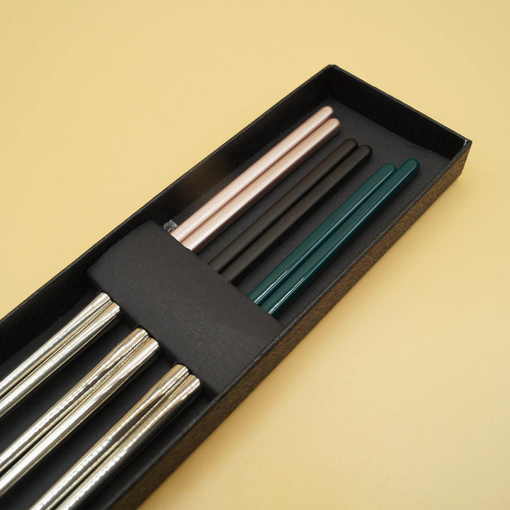 three sets of chopsticks lay in a box - one set with pink handles, one black, one dark green.