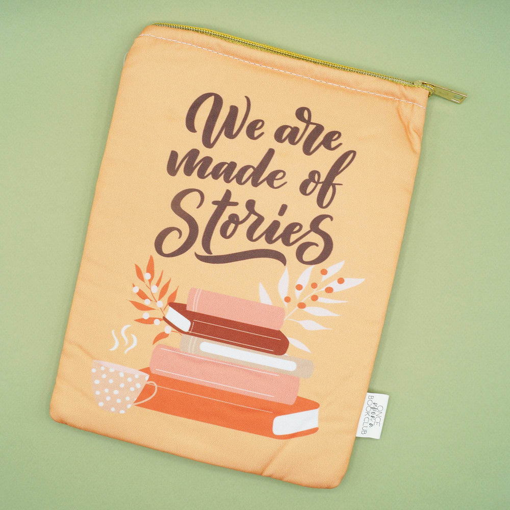 a peach-toned book sleeve with gold zipper closure. On the front is the quote "We are made of stories" above a stack of books