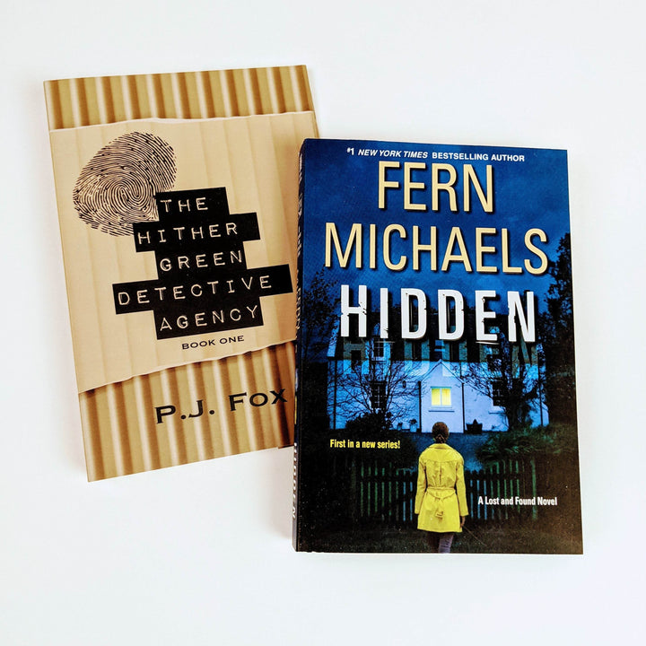 a light brown dust jacket with the title The Hither Green Detective Agency Book One by PJ Fox. A fingerprint is on the top left corner of the cover. The dust jacket is next to a mass market paperback edition of Hidden by Fern Michaels