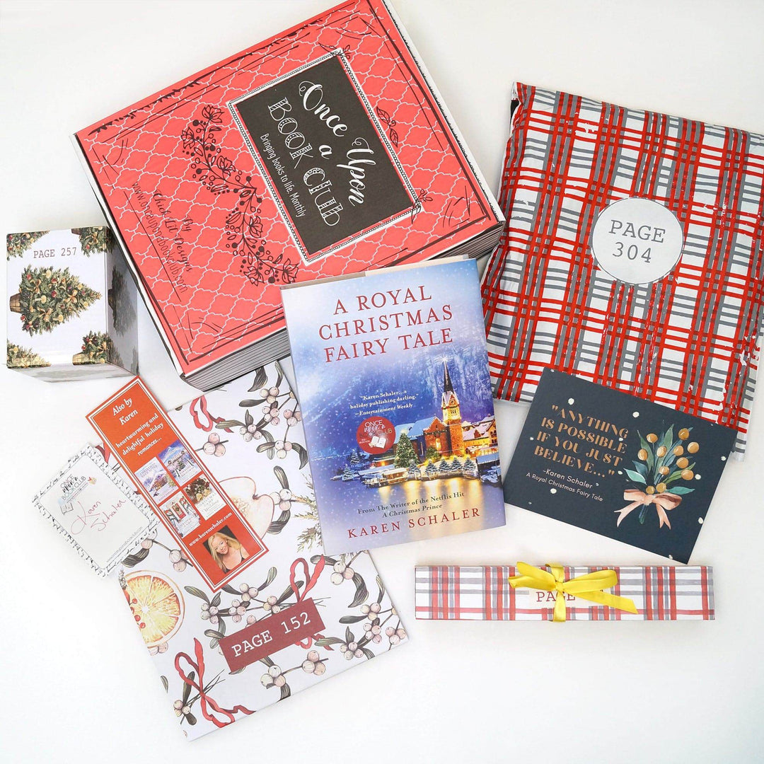 a hardcover edition of A Royal Christmas Fairy Tale leans against a red Once Upon a Book Club Christmas box. From left to right there is a square white box with Christmas tree pattern, signature card, bookmark, white folder, white box, quote card, and red and white polybag. The boxes, folder, and bag all have page numbers.