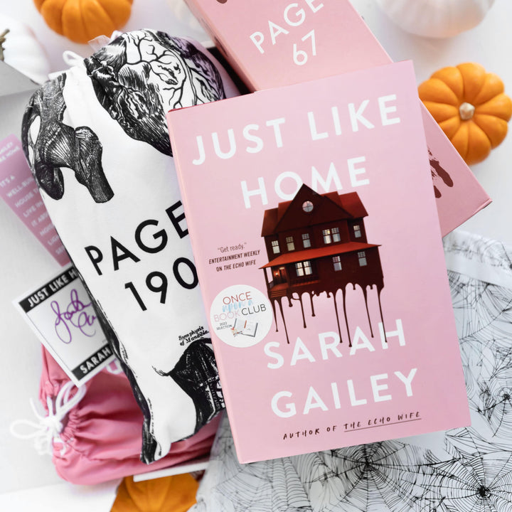 A hardcover edition of Just Like Home lays on a white and black drawstring bag, pink box, white polybag, pink drawstring bag, and assortment of paper items