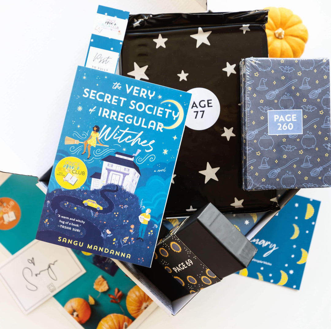 A paperback edition of The Very Secret Society of Irregular Witches lays on a black polybag with silver stars, assortment of paper items, black box, and blue box with witch-themed pattern