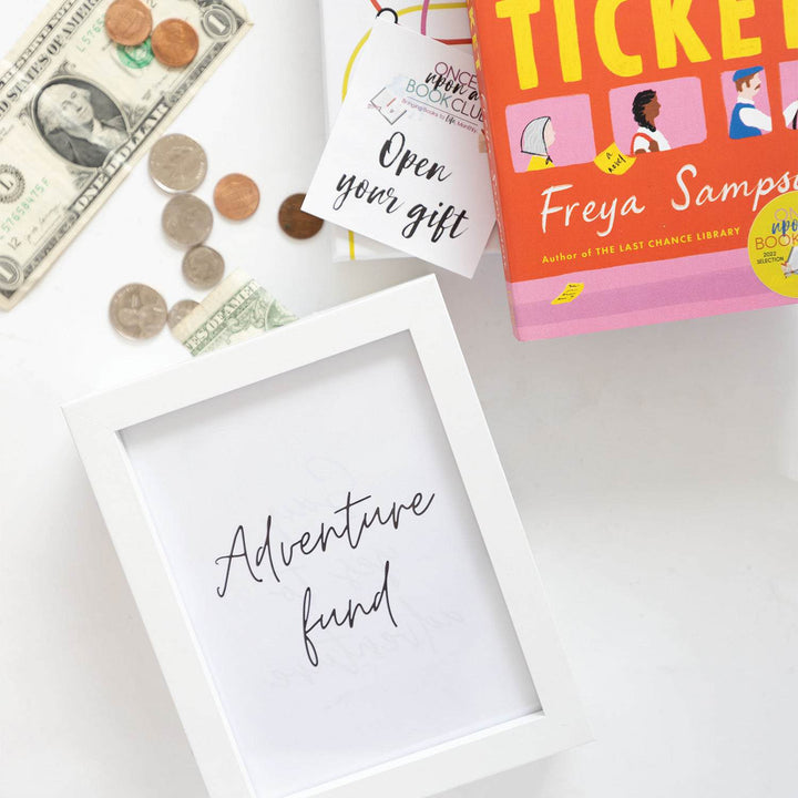 A white frame labeled Adventure Fund in black writing has money coming out of the top. It is next to a paperback edition of The Lost Ticket and an open your gift sticker.