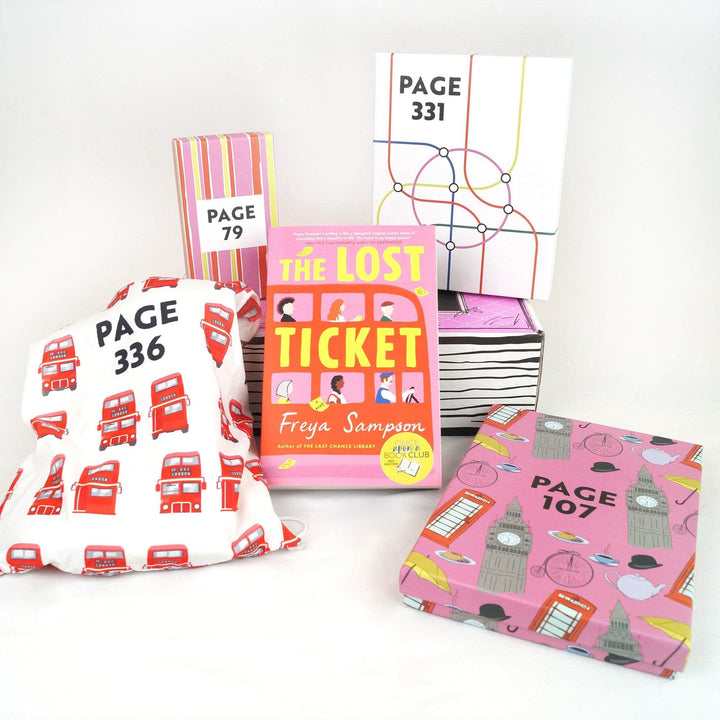 A pink box and white box are on a pink Once Upon a Book Club box. In front is a white drawstring bag, paperback edition of The Lost Ticket, and pink box. All boxes and bags are labeled with page numbers.