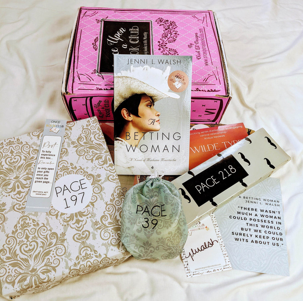 A paperback edition of The Betting Woman leans against a pink Once Upon a Book Club box. In front of the book and box are a gold and white box, bookmark, green drawstring bag, signature card, quote card, and white rectangular box with mustache pattern. The boxes and bags all have page numbers.