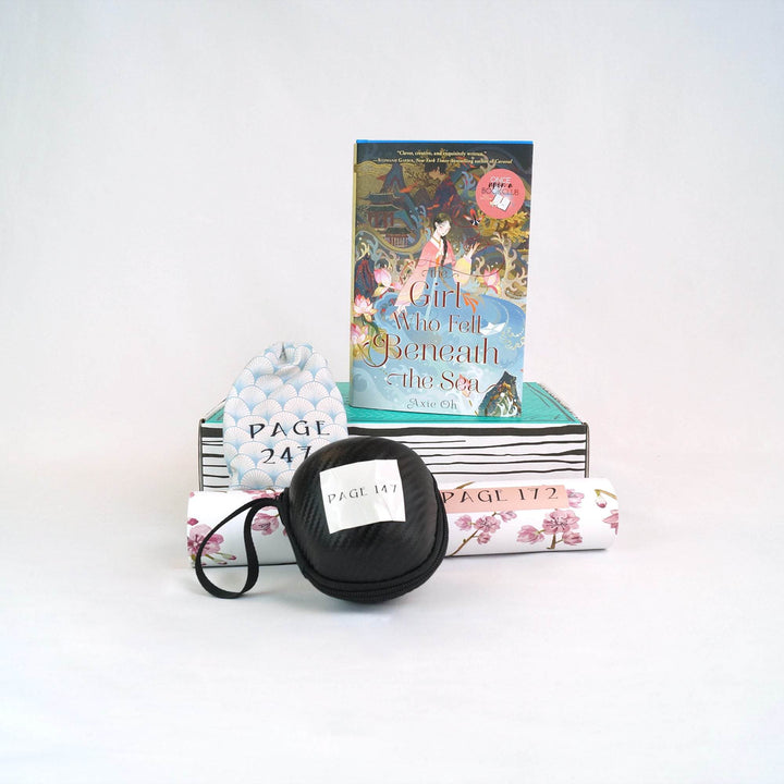 A hardcover edition of The Girl Who Fell Beneath the Sea stands on a green Once Upon a Book Club box. In front of the box are a blue and white drawstring bag, black ball, and white and pink tube. All gifts have page numbers on them.