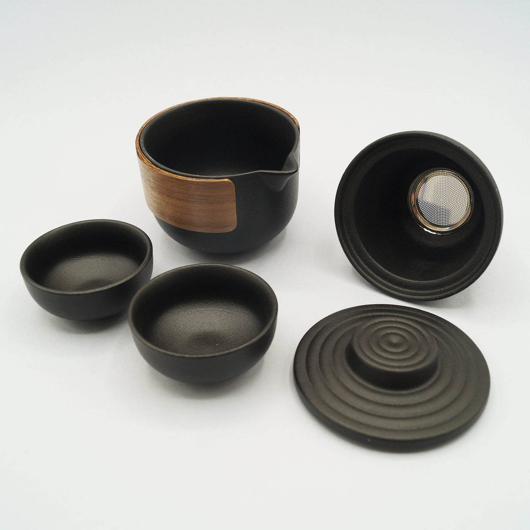 A black ceramic and wood tea set including one tea pot, one tea strainer, one lid, and two small tea cups. 