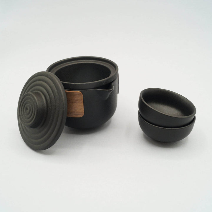 A black ceramic and wood tea set including one tea pot, one tea strainer, one lid, and two small tea cups stacked on top of each other