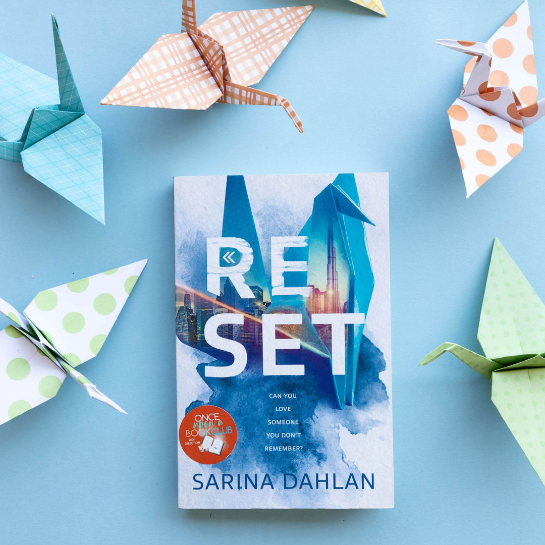 a paperback edition of Reset by Sarina Dahlan surrounded by paper cranes