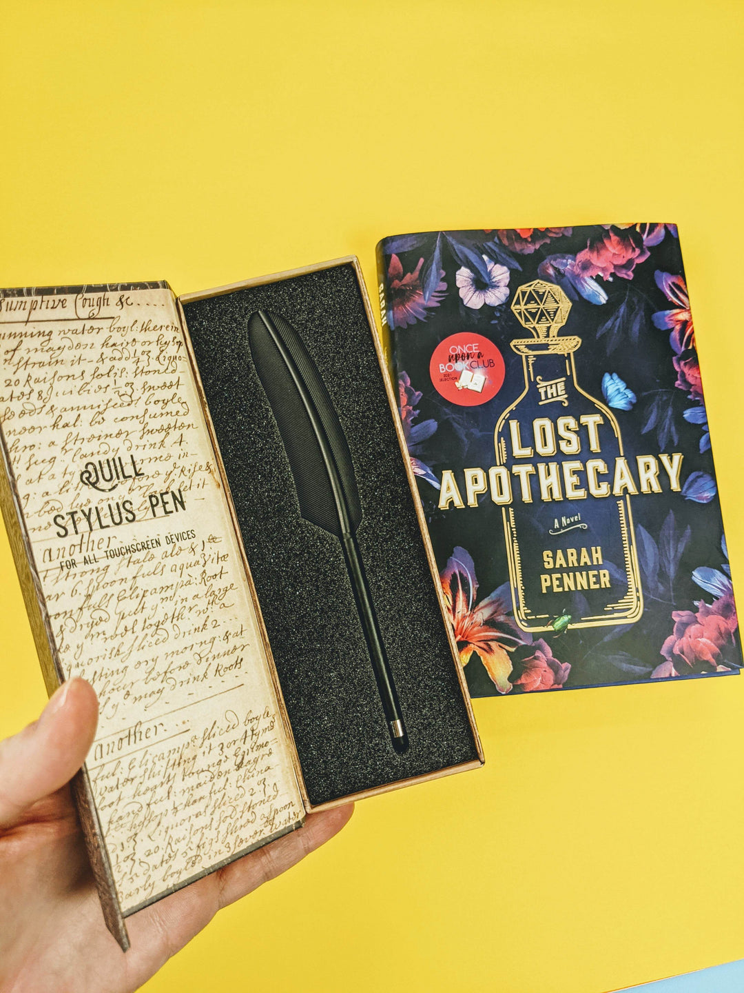 A white hand holds a box with a black quill feather stylus in it next to a hardcover edition of The Lost Apothecary