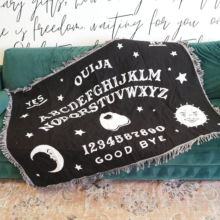a black woven blanket with a Ouija board pattern in white lays across a green couch
