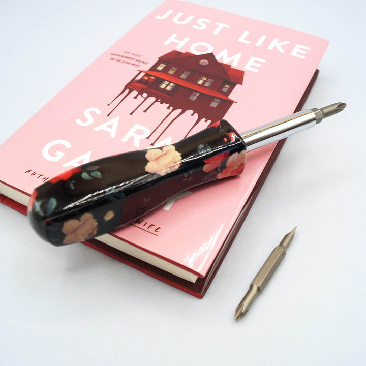 A screwdriver with a black handle with red flowers is laying on a hardcover edition of Just Like Home