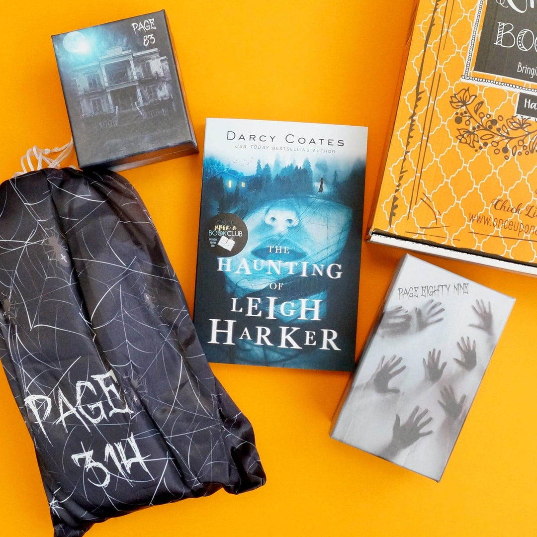 A paperback edition of The Haunting of Leigh Harker is centered in the photo. Surrounding the book are a black drawstring bag with a spiderweb pattern, black square box, white box with handprints on it, and an orange Once Upon a Book Club box