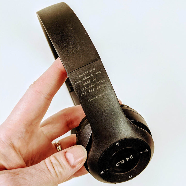 a white hand holds black over-the-ear wireless headphones. A quote is printed on the them that says "Whatever our souls are made of, his and mine are the same"
