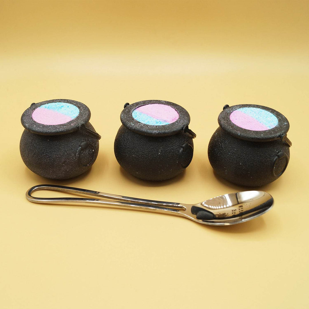 3 black cauldron-shaped bath bombs stand in a line behind a silver spoon