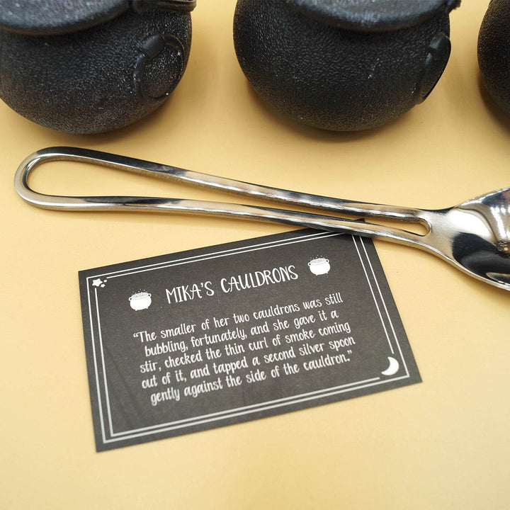 black cauldron-shaped bath bombs stand behind a silver spoon and a black card that says Mika's Cauldrons