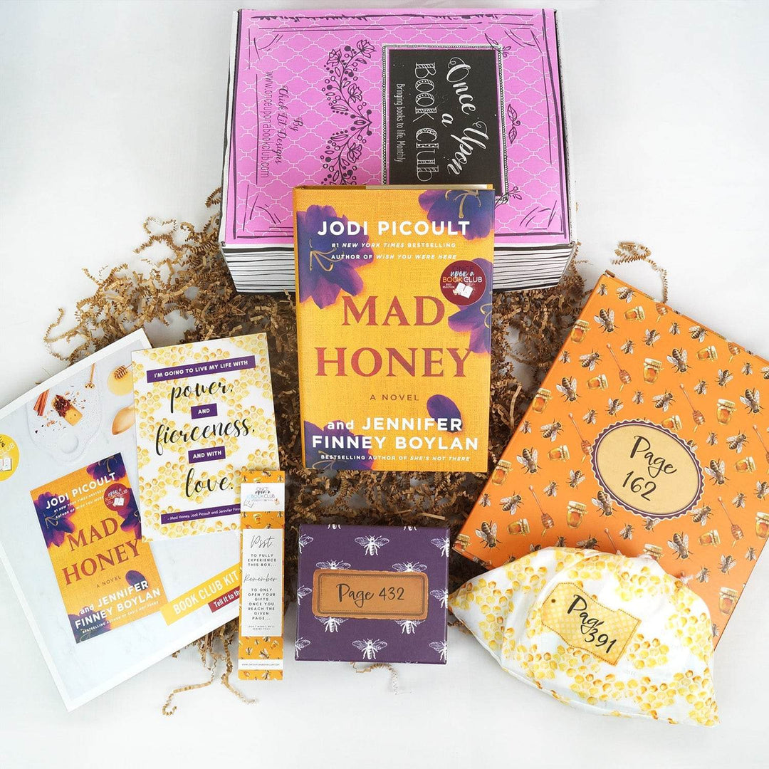 A hardcover edition of Mad Honey leans against a pink Once Upon a Book Club box. In front are a bookclub kit, quote card, bookmark, purple box, orange folder, and yellow and white drawstring bag. The bag, box, and folder are all labeled with page numbers