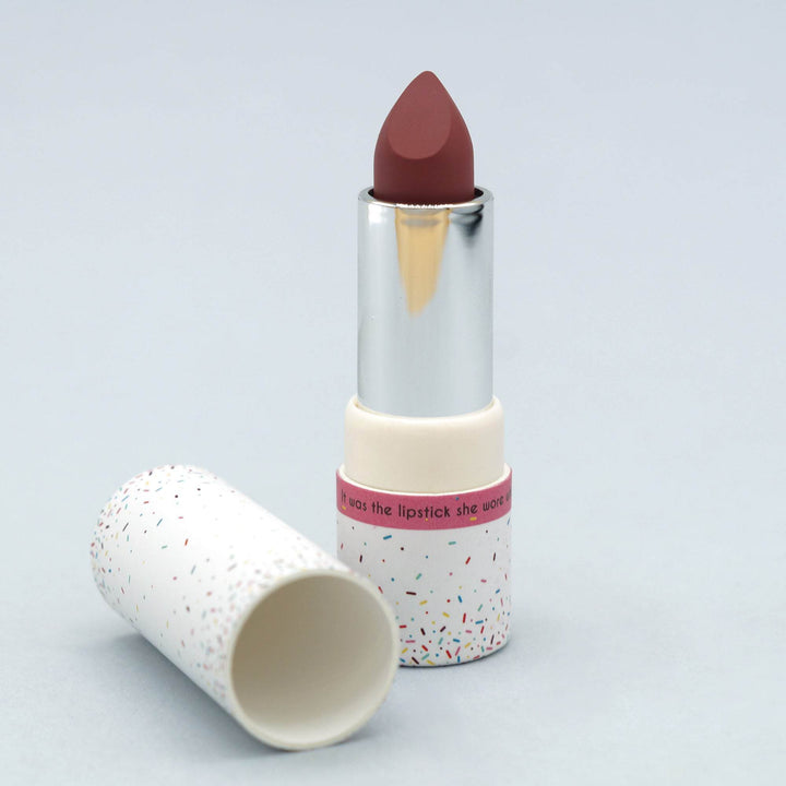 A white lipstick tube with multicolored sprinkles pattern. The lipstick is open with the lid beside it and shows a berry red color.