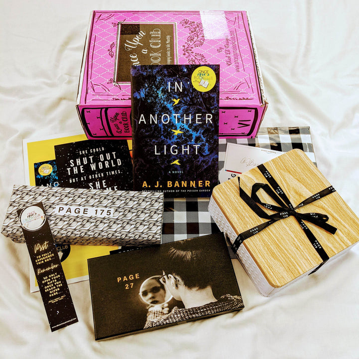 A paperback edition of In Another Light leans against a pink Once Upon a Book Club box. In front of the book are a black and white rectangular box, black box, white box with wooden lid, and assortment of paper items. The boxes and bags all have page numbers.