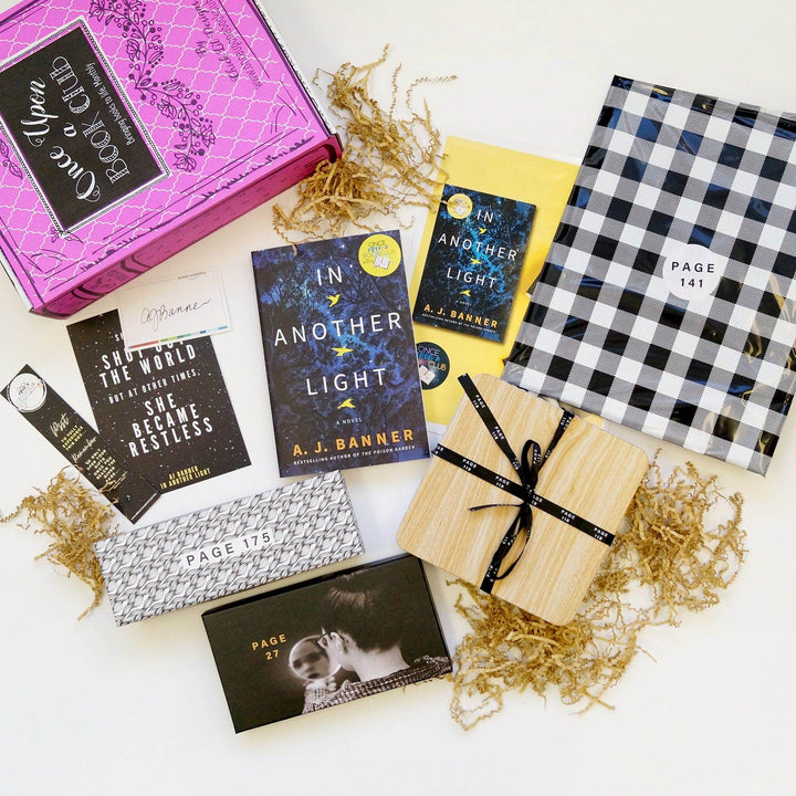 A pink Once Upon a Book Club box is in the top left corner. In front of the box are (from left to right) a bookmark, quote card, signature card, black and white box, black box, paperback edition of In Another Light, bookclub kit, box with wooden lid, and a black and white checkered polybag. The boxes and bags all have page numbers on them.