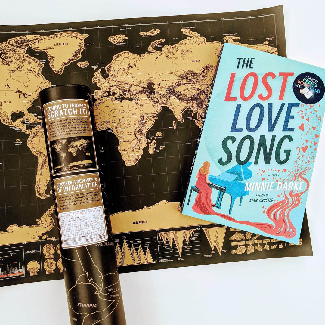 A paperback edition of The Lost Love Song lays on a black and gold scratch off map of the world.