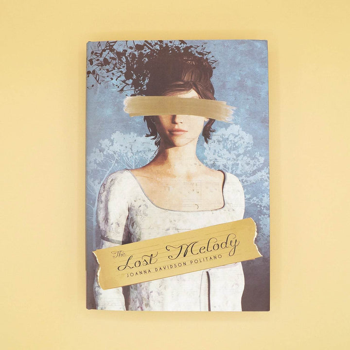 The front dustjacket of a hardcover special edition of The Lost Melody. The cover shows a white woman wearing a white dress with a gold brushstroke over her face and her dark hair is floating away in pieces.