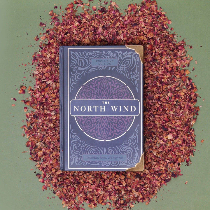 A hardcover special edition of The North Wind by Alexandria Warwick with gold metal corners laying on a pile of rose petals