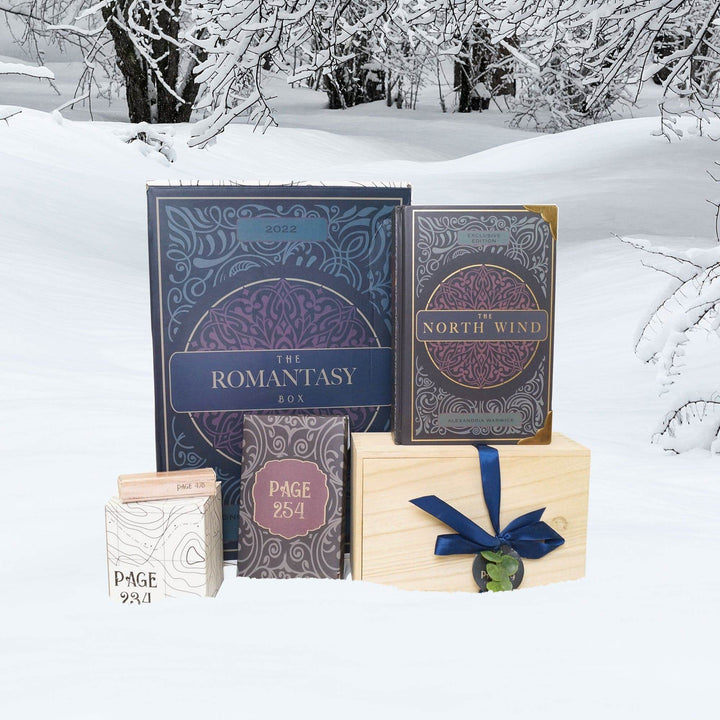 A hardcover special edition of The North Wind with metal corners is on top of a wooden box next to a Once Upon a Book Club Romantasy box, a black box, small beige box, and white box. The boxes are all labeled with page numbers.