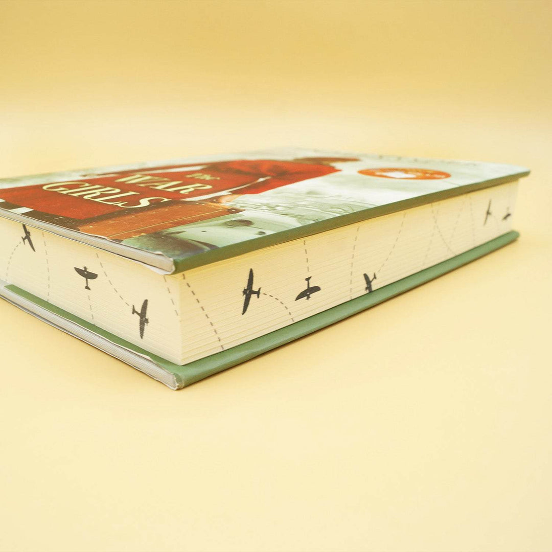 A hardcover special edition of The War Girls lays on its side. The page edges show black airplanes flying in different directions.
