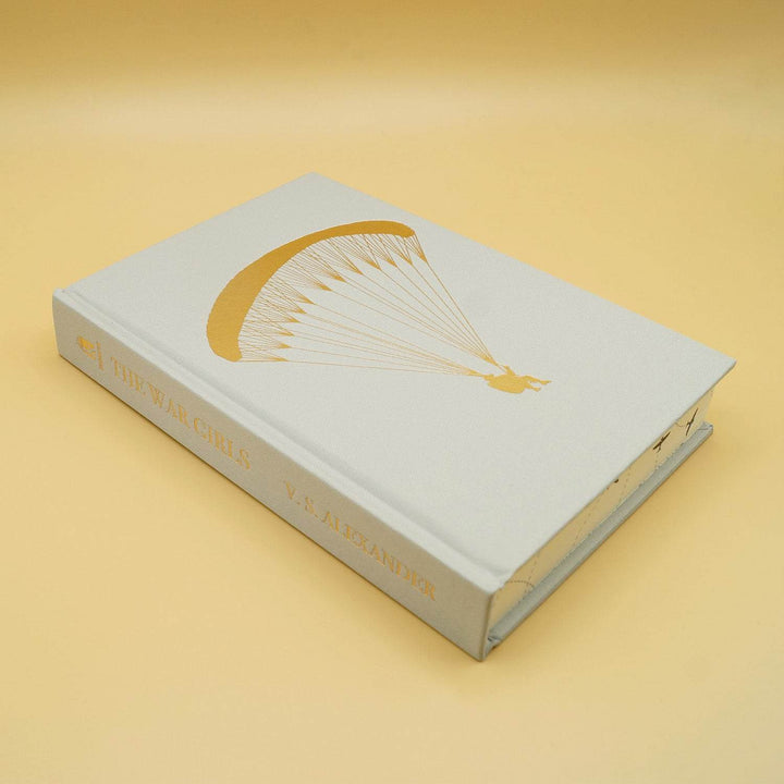 The hardcase of a special edition of The War Girls. The book is white with a gold image of someone flying down in a parachute