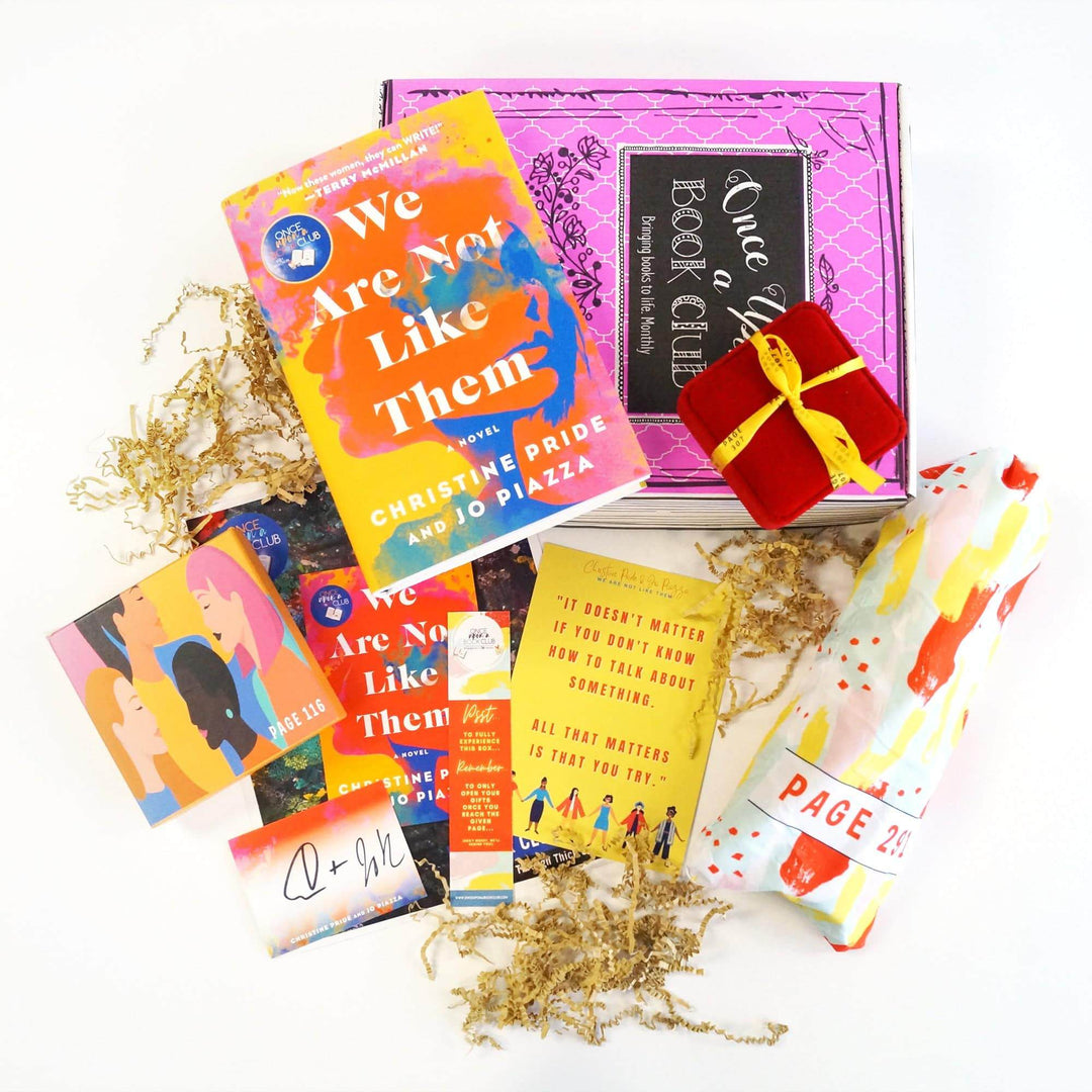 A hardcover edition of We Are Not Like Them and a red square box are on top of a pink Once Upon a Book Club box. In front of the box are a yellow square box, bookclub kit, signature card, bookmark, quote card, and white drawstring bag. The boxes and bags all have page numbers on them.