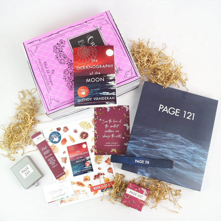 A paperback edition of The Oceanography of the Moon is on a pink Once Upon a Book Club box. In front of the box are a silver square box, bookmark, signature card, bookclub kit, quote card, large blue box, long blue box, and pink square box. The boxes and bags all have page numbers.