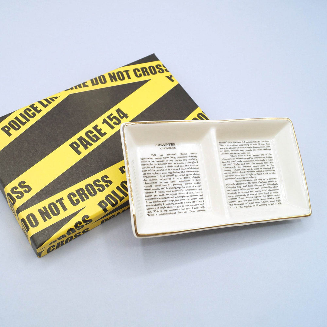 a white ceramic and gold painted ring dish with two sections, featuring the first page of Moby Dick by Herman Melville. The ring dish is leaning against a box lid that is black with crime scene tape across it, labeled with a page number