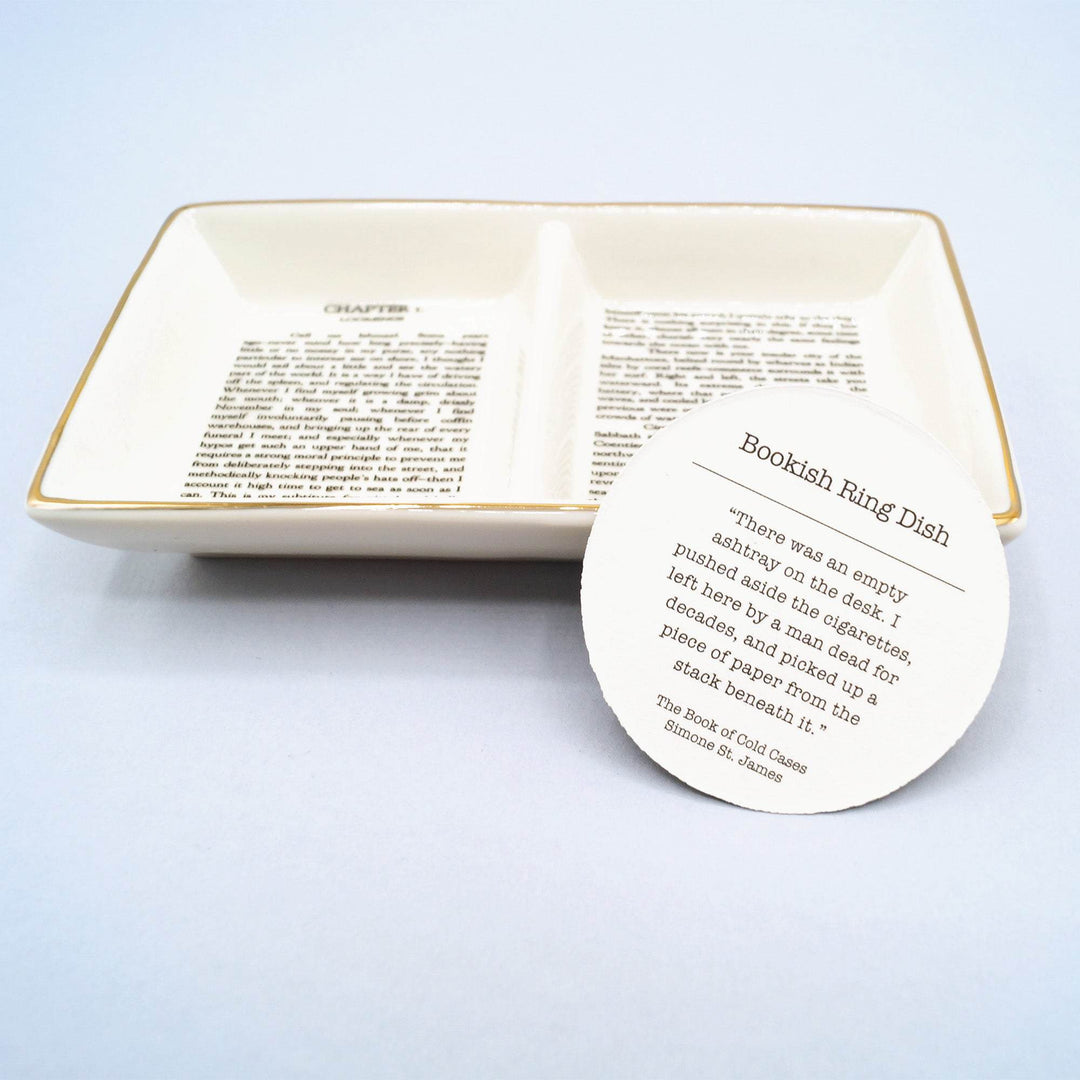 a white ceramic and gold painted ring dish with two sections, featuring the first page of Moby Dick by Herman Melville. A circular information card leans against it.