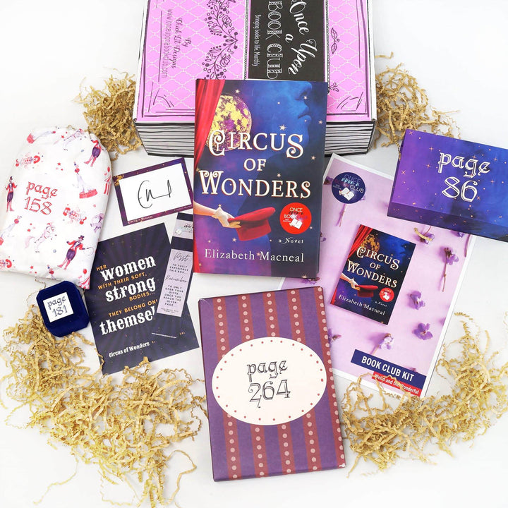 A hardcover edition of Circus of Wonders is leanings against a pink Once Upon a Book Club box. In front of the box are a white drawstring bag, small blue felt ring box, signature card, quote card, bookmark, purple and red box, bookclub kit, and purple box. The boxes and bags all have page numbers.