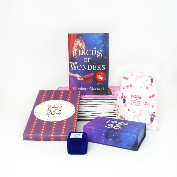A hardcover edition of Circus of Wonders is standing on a pink Once Upon a Book Club box. In front of the box are a purple and red box, blue felt ring box, purple and blue box, and white drawstring bag. The boxes and bags all have page numbers.