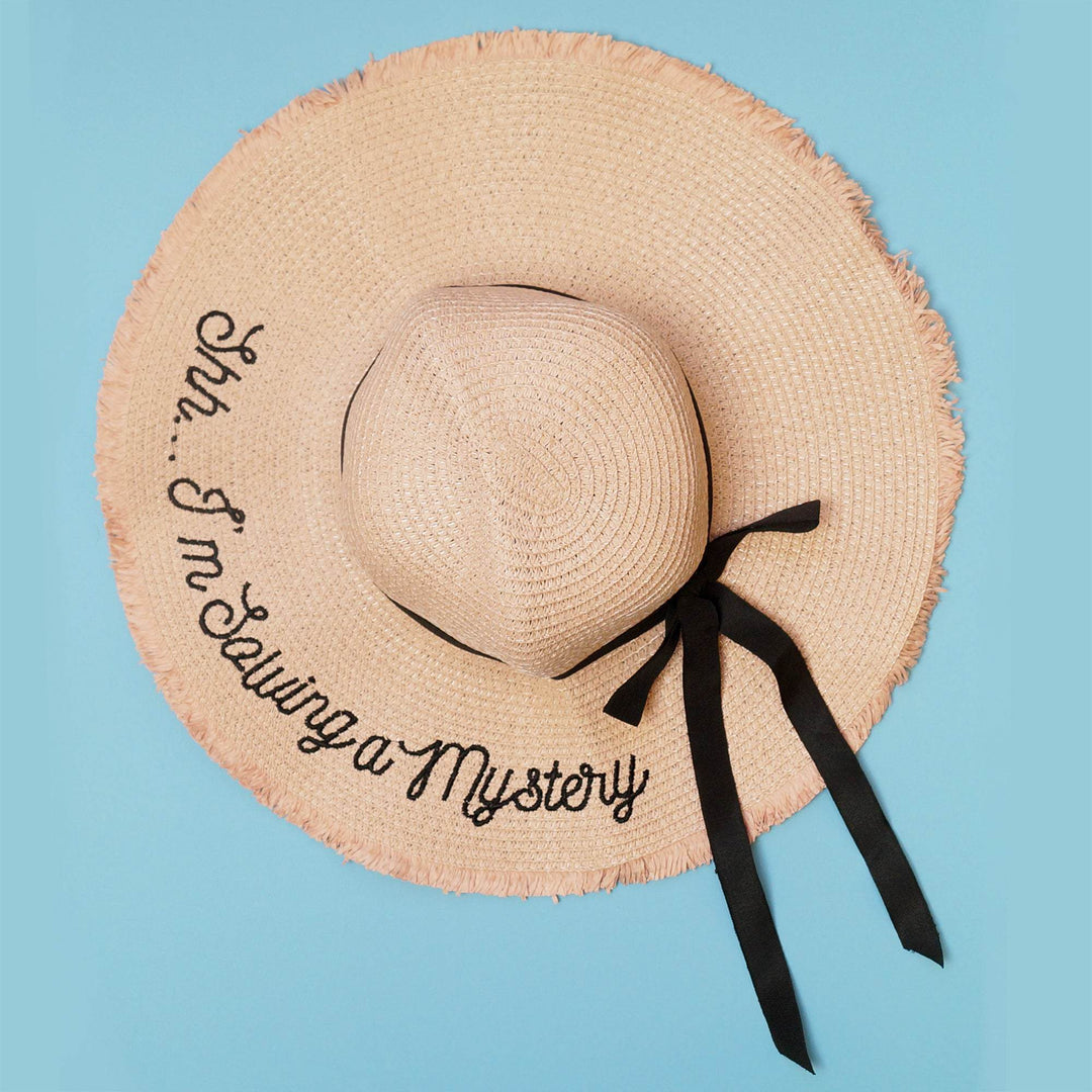 a blush pink sun hat featuring the words "Shh...I'm Solving a Mystery" embroidered in black thread, finished with a black ribbon