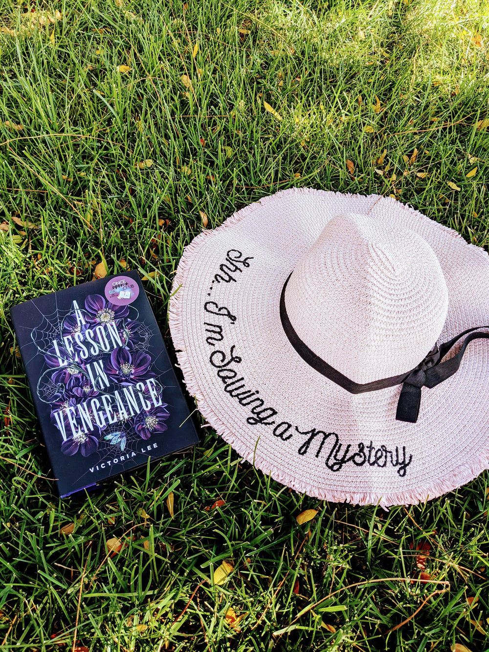 A hardcover edition of A Lesson in Vengeance is laying in the grass next to a blush pink sun hat featuring the words "Shh...I'm Solving a Mystery" embroidered in black thread, finished with a black ribbon