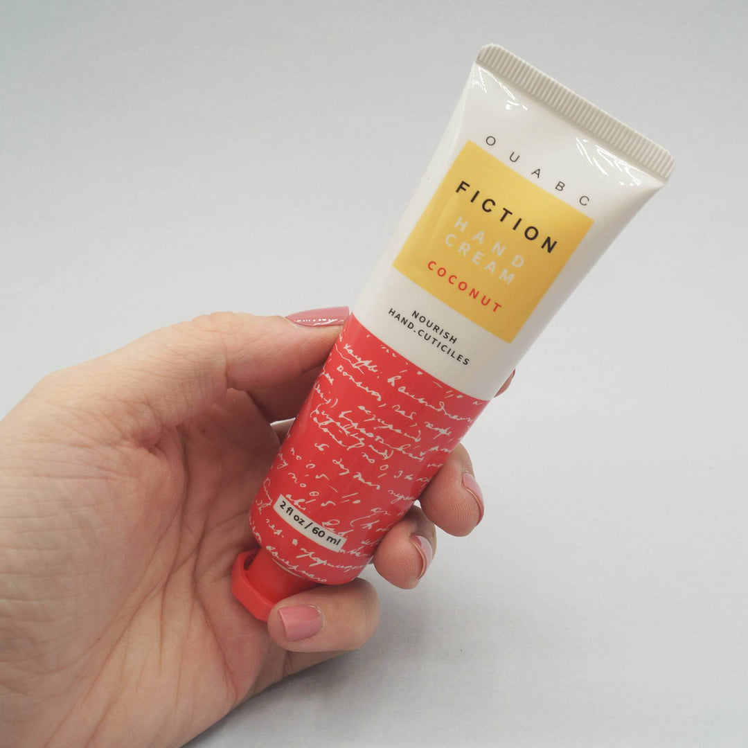 a white hand holds a 2 fl oz red-orange and white tube of "OUABC Fiction Hand Lotion - Coconut" 