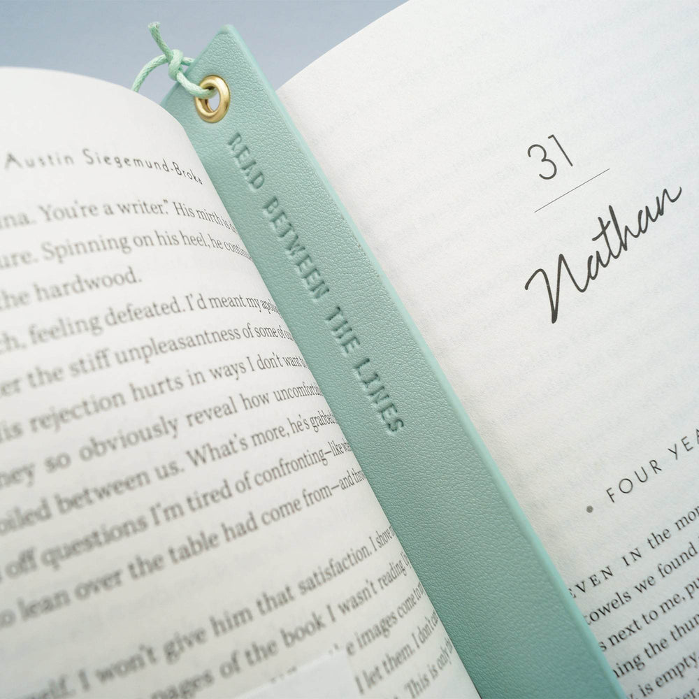 a seafoam green leather bookmark with the quote "Read between the lines" lays in an open book