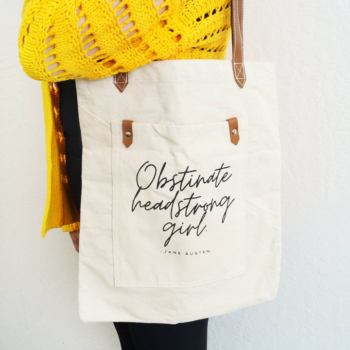 A woman wearing a yellow sweater carries a beige canvas tote bag printed with the Jane Austen's quote, "Obstinate headstrong girl" 